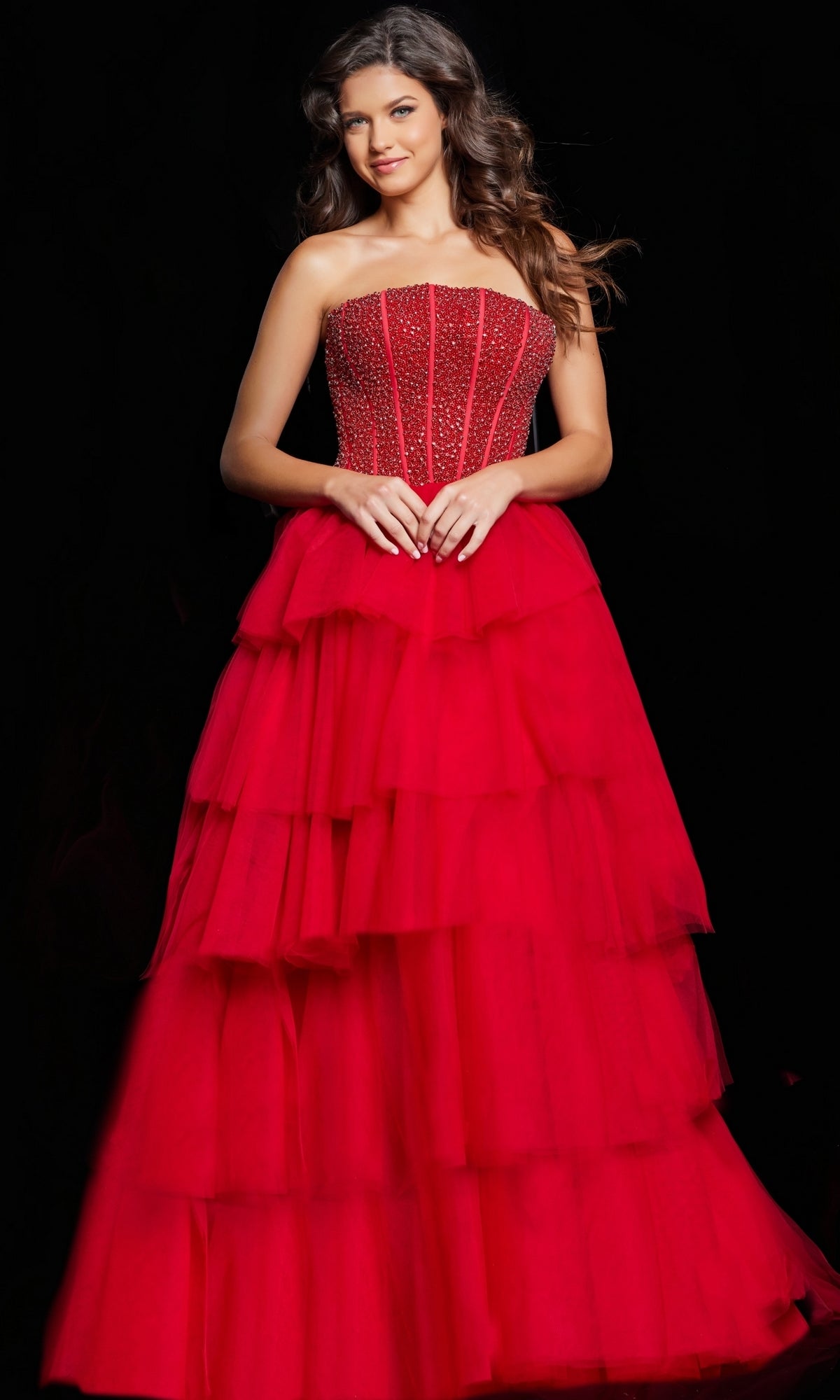 A woman stands against a black background wearing the Long Prom Dress 37210 by Jovani, a strapless tulle ball gown featuring an embellished corset bodice and layered skirt.